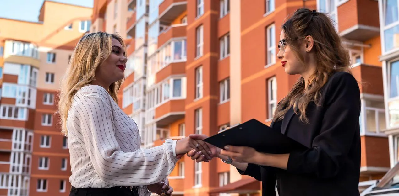Two women, one a buyer's agent, shaking hands in front of an apartment building, sealing a successful deal.